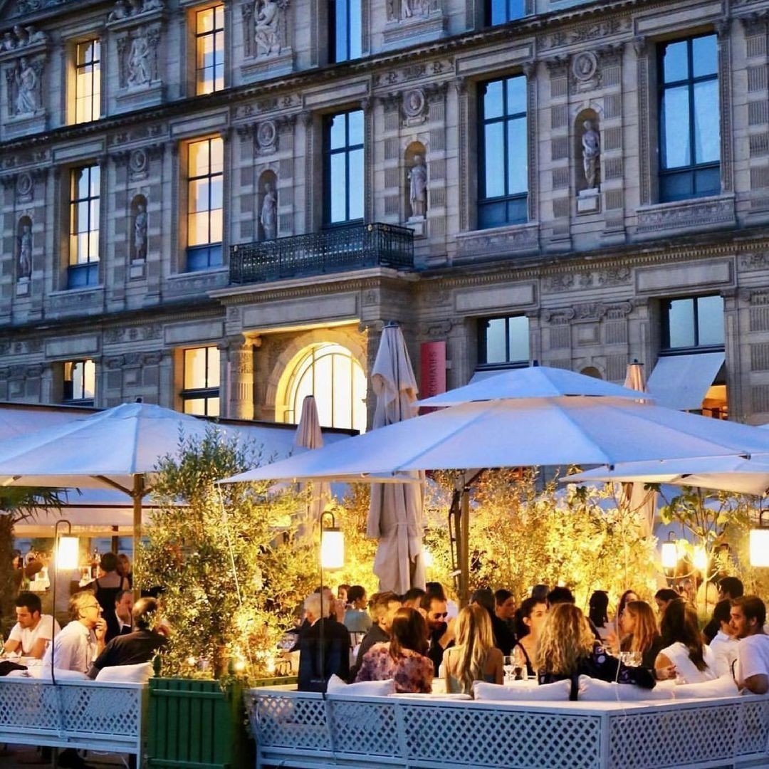 Outdoor evening terrace party with people under white umbrellas and string lights, elegant historic building backdrop