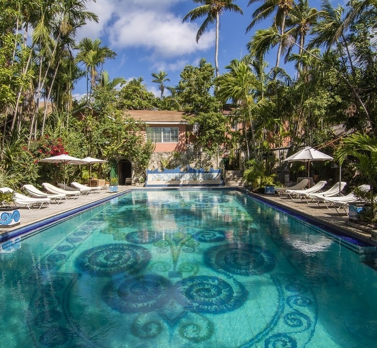 Luxurious swimming pool with turquoise water and decorative floor patterns, flanked by lounge chairs and umbrellas, surrounded by palm trees and a terracotta villa.