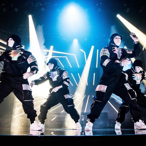 K-pop group performing on stage in coordinated black outfits with dramatic blue lighting and pyrotechnics