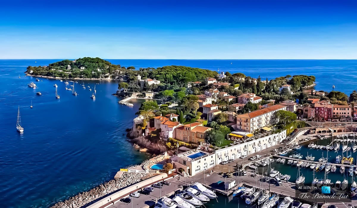 Aerial view of a Mediterranean coastal town with colorful buildings, marina, and sailing boats surrounded by turquoise water