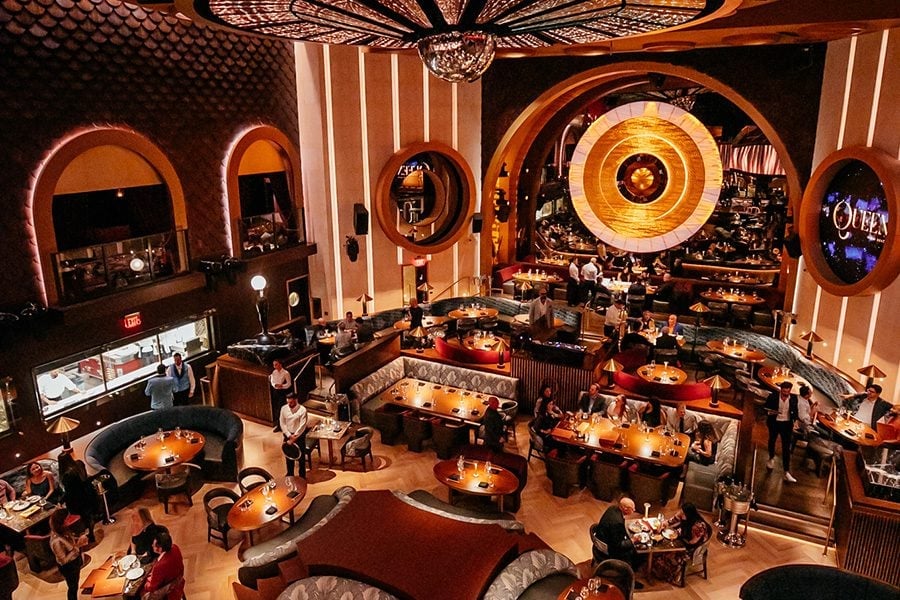 Overhead view of an upscale restaurant interior with warm orange and gold lighting, circular tables, arched alcoves, and a large illuminated circular design on the ceiling