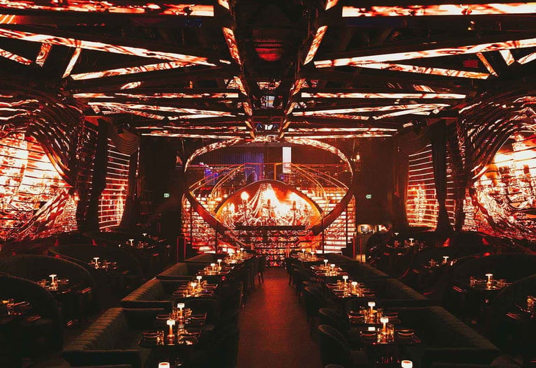 Symmetrical nightclub interior with golden geometric lighting, rows of tables on both sides, and a stage glowing in warm orange and red tones