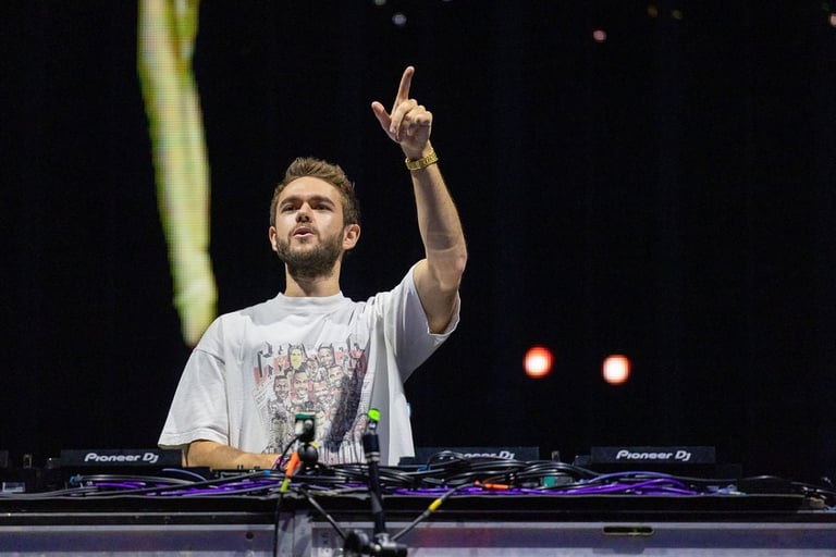 DJ performing at turntables on stage, pointing upward to the crowd with one hand, wearing a light gray t-shirt against a dark background with stage lighting