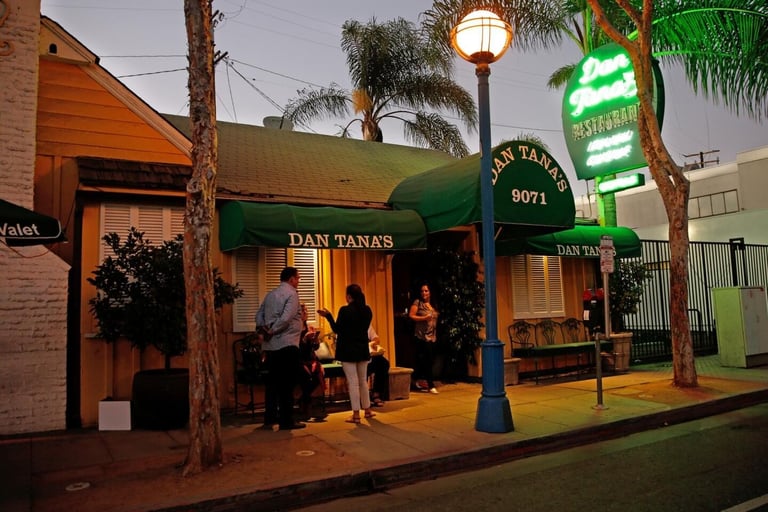 Exterior of Dan Tanas restaurant at night with illuminated neon signage, palm trees, and customers standing outside the entrance
