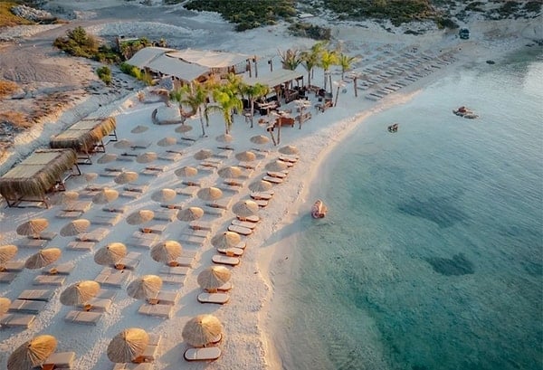 Aerial view of a beach resort with tan umbrellas lining the sandy shore and turquoise water with boats