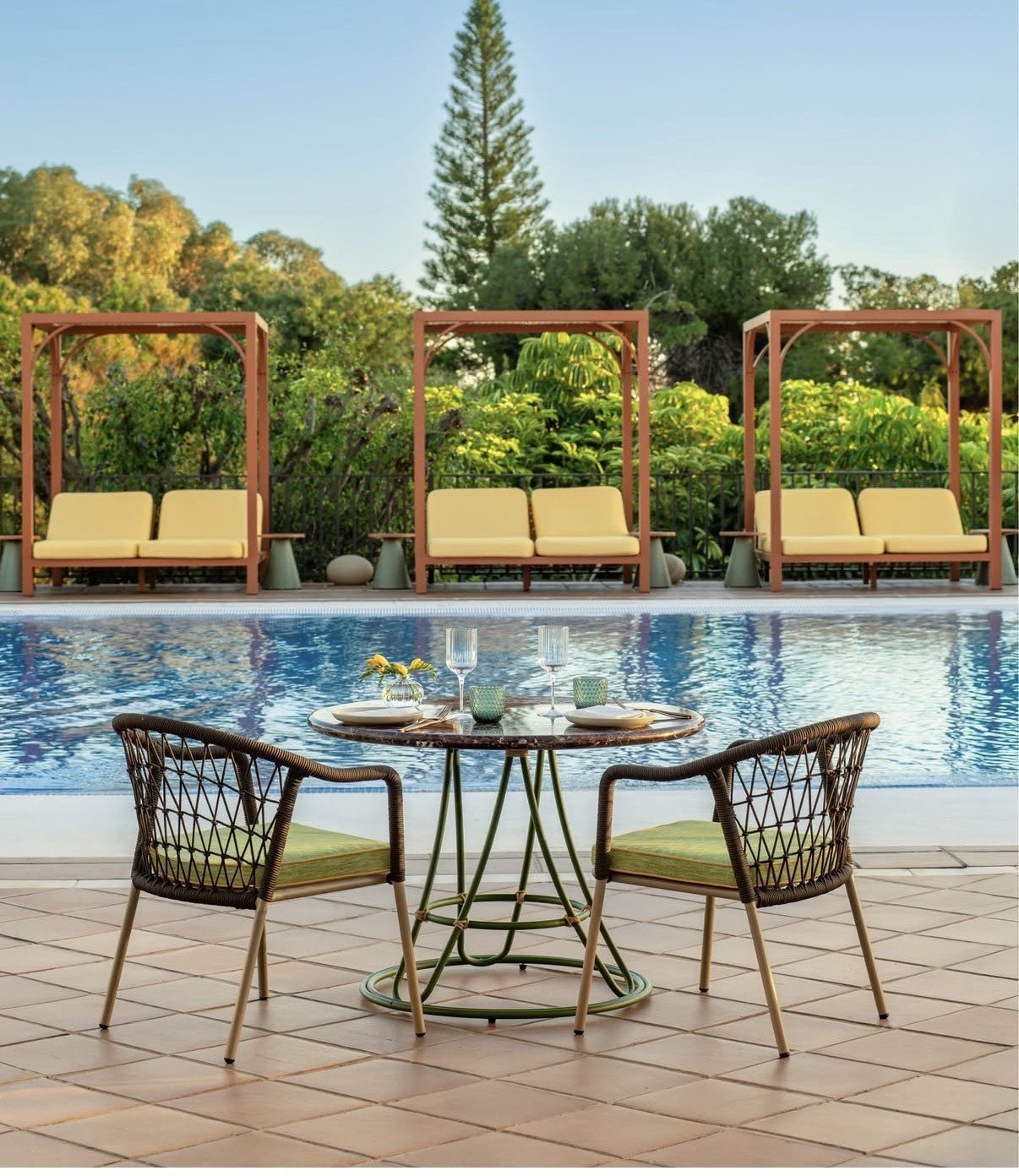 Modern outdoor pool area with woven lounge chairs, round glass table, wooden pergolas, and lush green landscaping