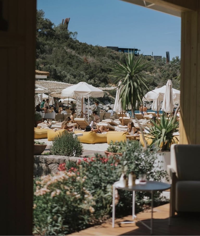 Sunny Mediterranean outdoor lounge with white umbrellas, yellow seating areas, palm trees, and hillside view framed through an open doorway