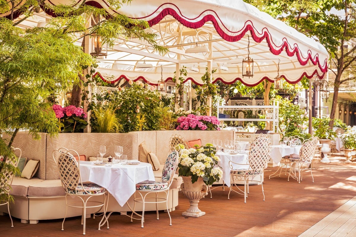 Elegant outdoor patio dining area with red and white striped awning, white tablecloths, beige chairs, and flowering plants.