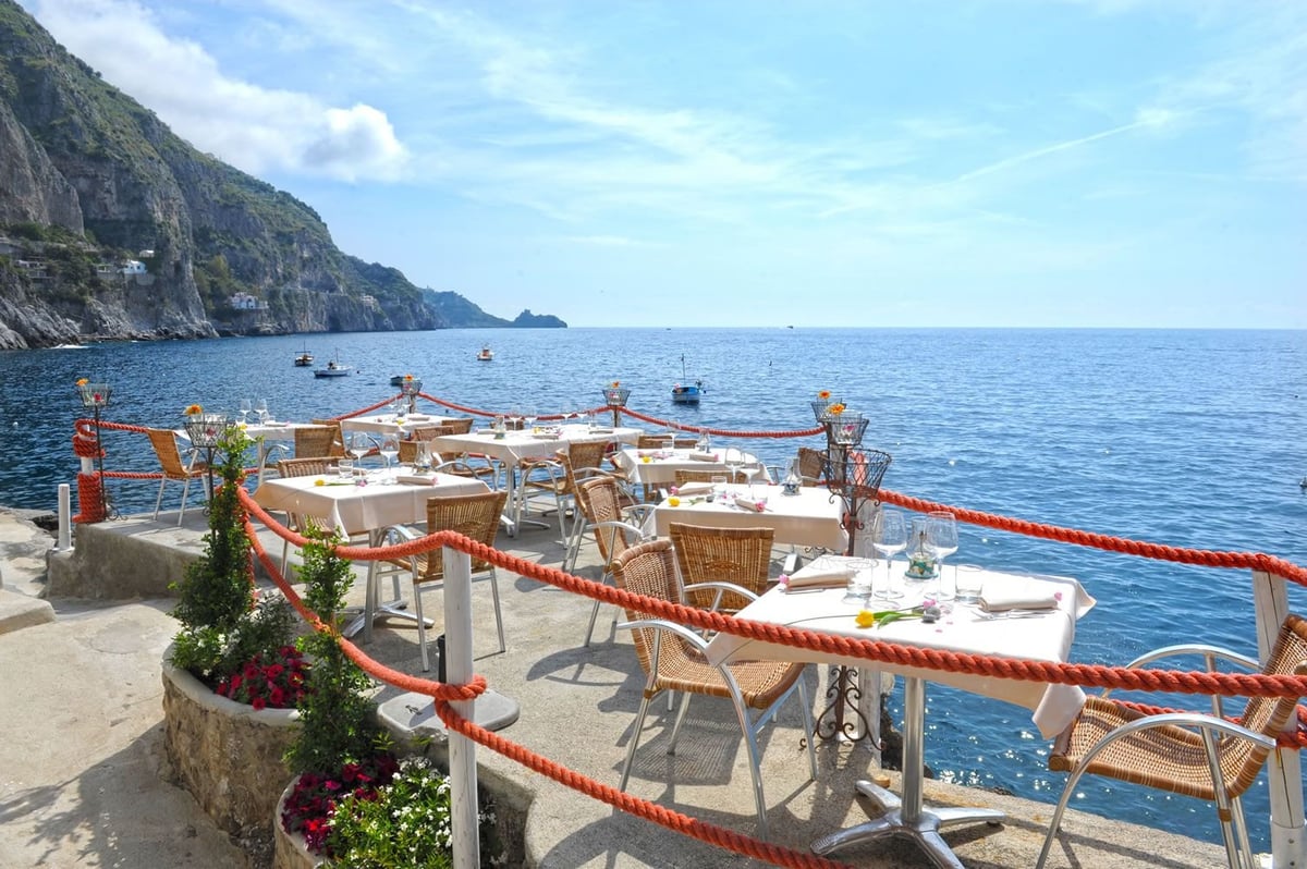 Seaside restaurant terrace with wooden tables and chairs overlooking blue Mediterranean sea and rocky cliffs