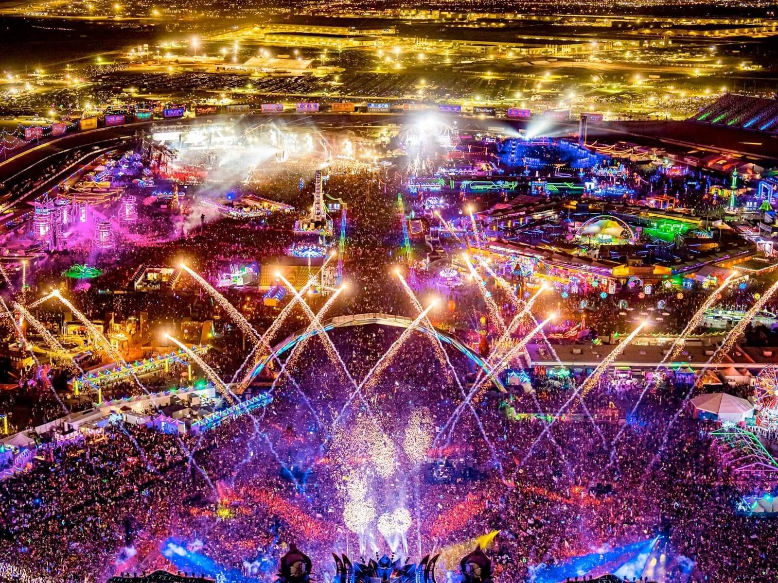 Aerial view of a large outdoor music festival at night with colorful stage lights in pink, blue, and gold illuminating a massive crowd.