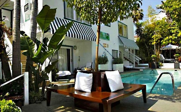 Modern poolside lounge area with wooden daybed and white pillows, tropical plants, white villa, and swimming pool in background