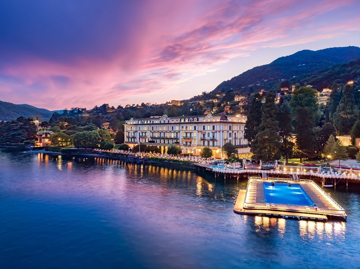 Luxury waterfront villa at dusk with floating pool on lake, surrounded by illuminated mountains and buildings reflected in calm blue water