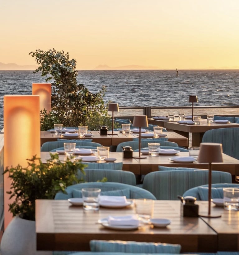 Waterfront restaurant terrace at sunset with set tables, upholstered chairs, and sea views