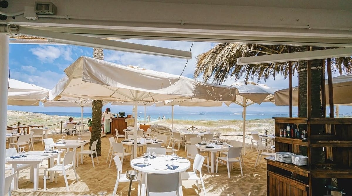 Beachfront restaurant dining area with shaded umbrellas, white tables and chairs overlooking turquoise water and sandy beach