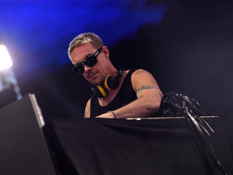 DJ performing at turntables with arms raised triumphantly in front of illuminated green LED screens