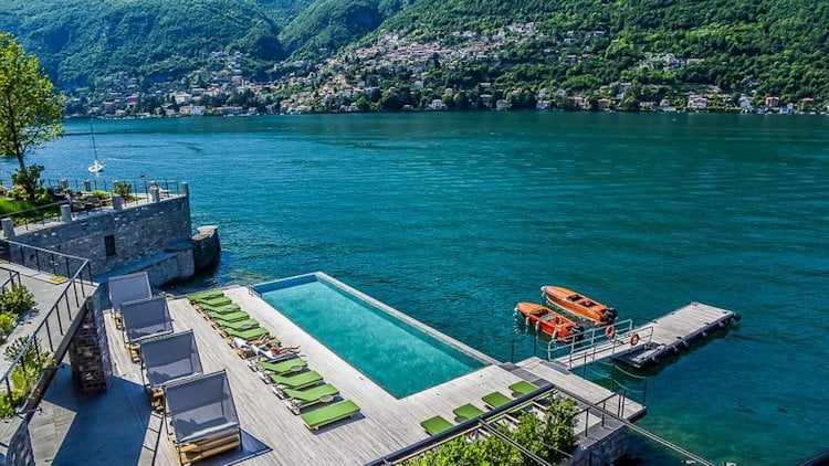 Luxury lakeside pool with sunbeds overlooking turquoise water, mountain village, and wooden boat dock