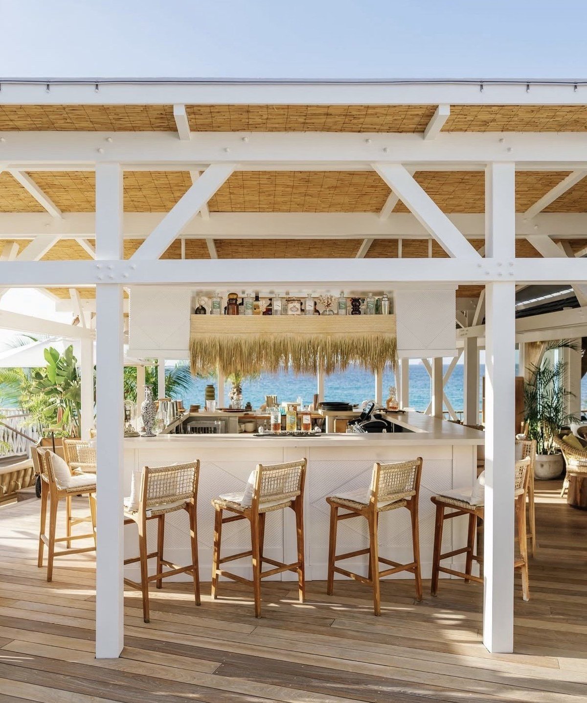 Open-air beachfront bar with white pergola, wooden bar stools, and ocean views under a thatched roof