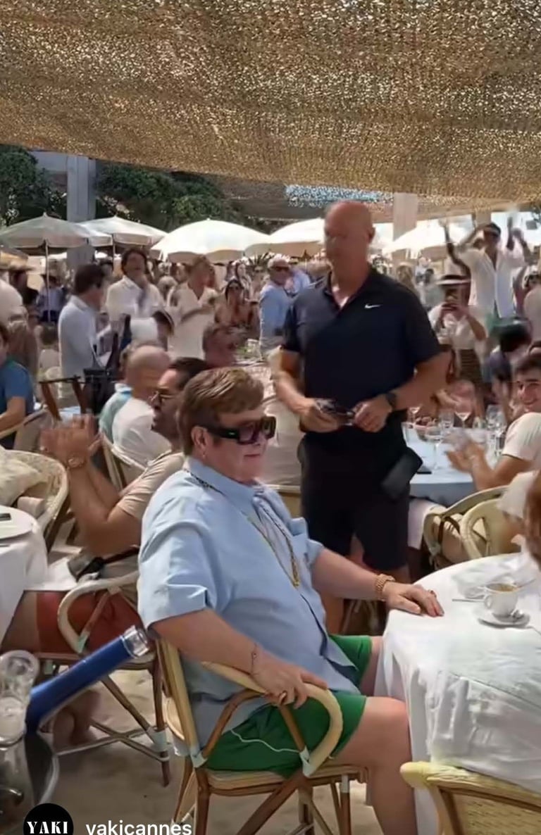 Crowded outdoor dining event under shade canopy with guests seated at tables and a man in black Nike shirt standing