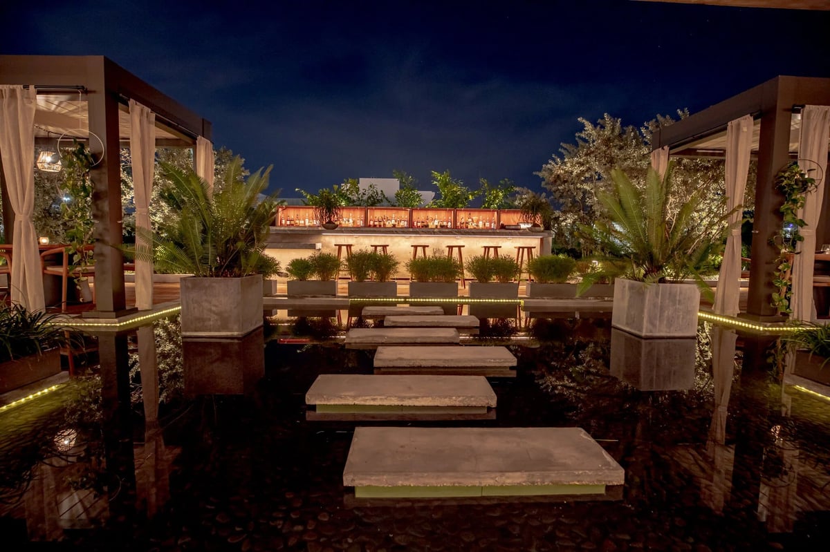 Illuminated pathway with stepping stones at night, flanked by potted plants and pergolas, overlooking a lit courtyard building
