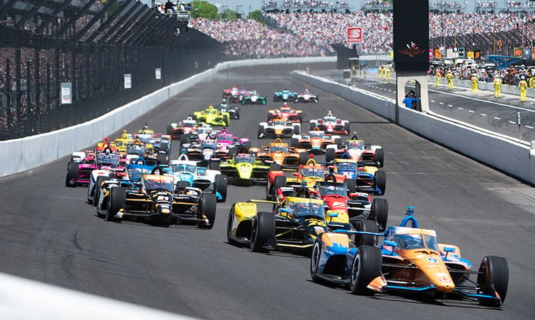 Formula racing cars competing on an oval track with crowded grandstands in the background during a professional motorsports event.