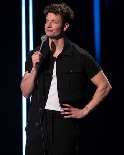 Man performing on stage holding a microphone, wearing a black shirt over a white t-shirt against a dark background with blue lighting