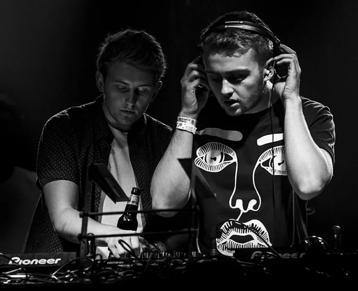 Two male DJs performing at a turntable setup with dramatic cloudy sky backdrop during an electronic music performance