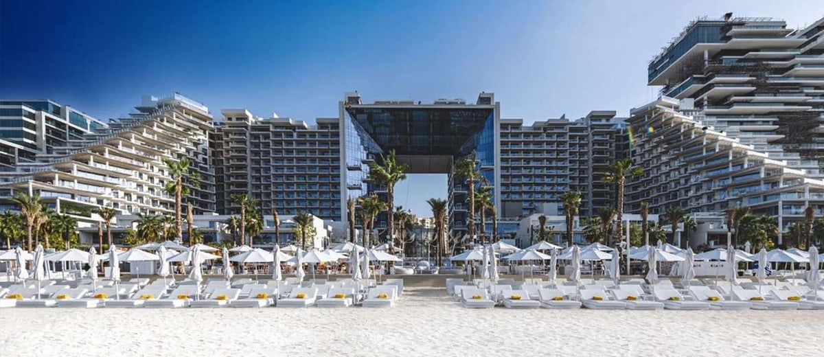 Luxury beachfront resort with white sun loungers and umbrellas on sandy beach, tall residential towers and palm trees under blue sky