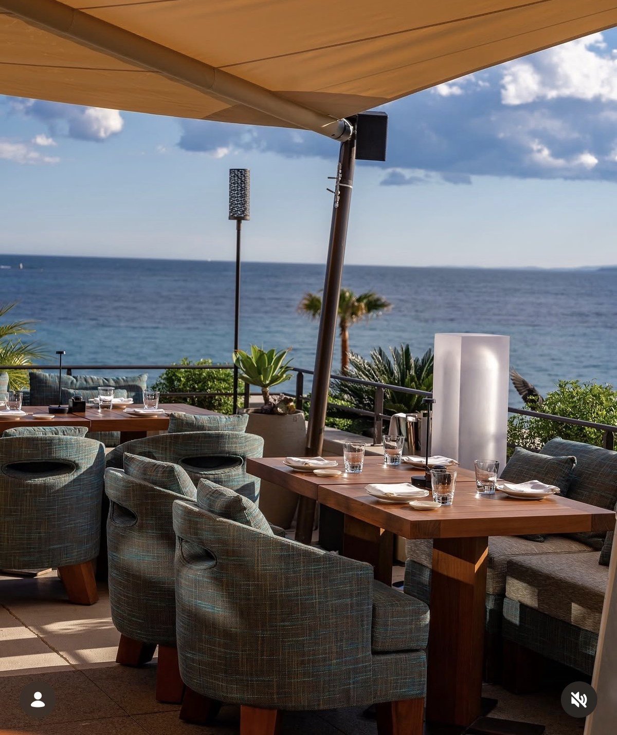 Outdoor oceanfront dining area with gray upholstered chairs under tan awning, wooden table, and sea view