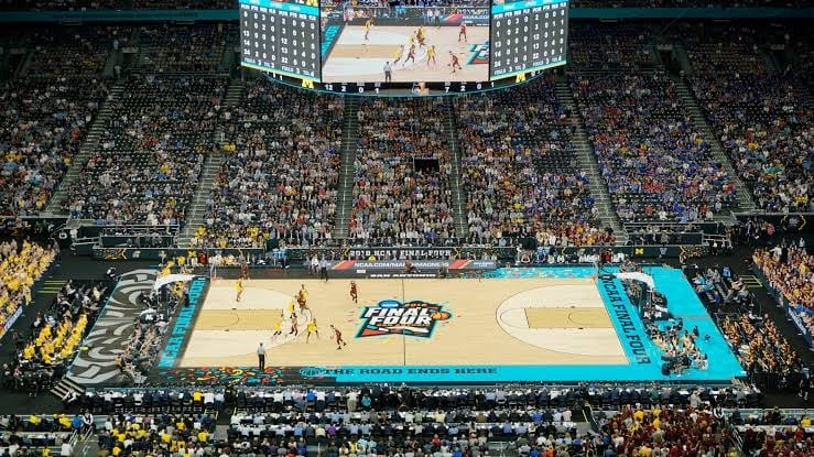 Packed basketball arena with large crowd in stands, viewing game on court below with blue sidelines and scoreboard overhead