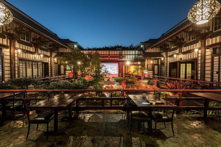 Elegant courtyard at night with traditional wooden buildings, seating area, and illuminated landscaping under a clear sky