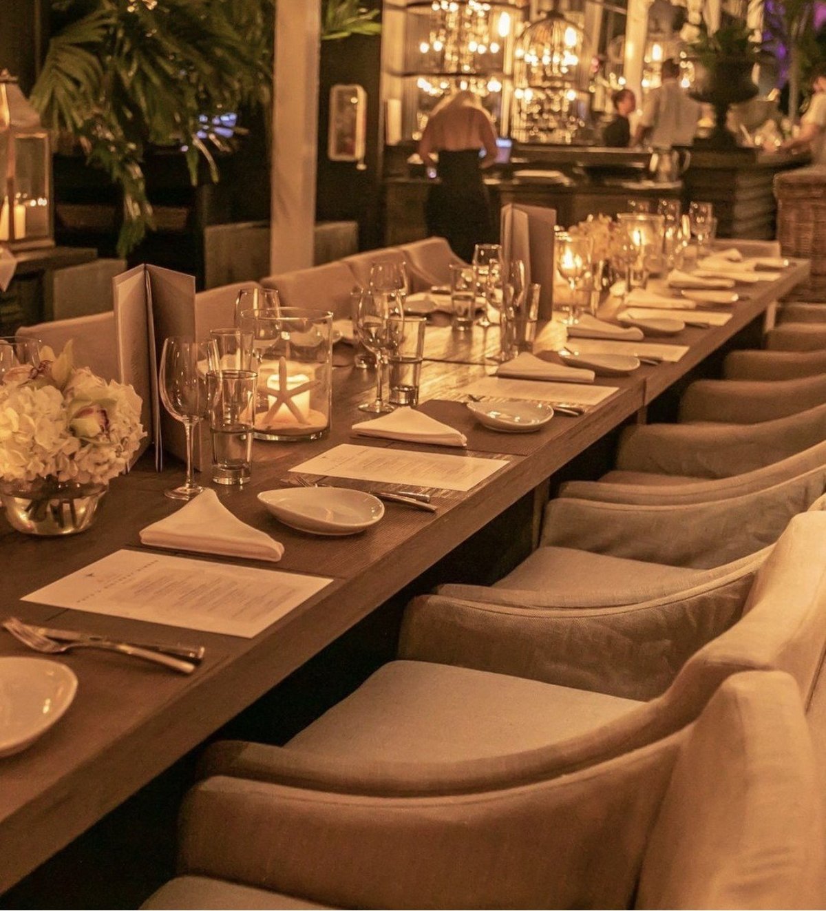 Elegant formal dining room with long wooden table, upholstered beige chairs, place settings, wine glasses, and warm ambient lighting