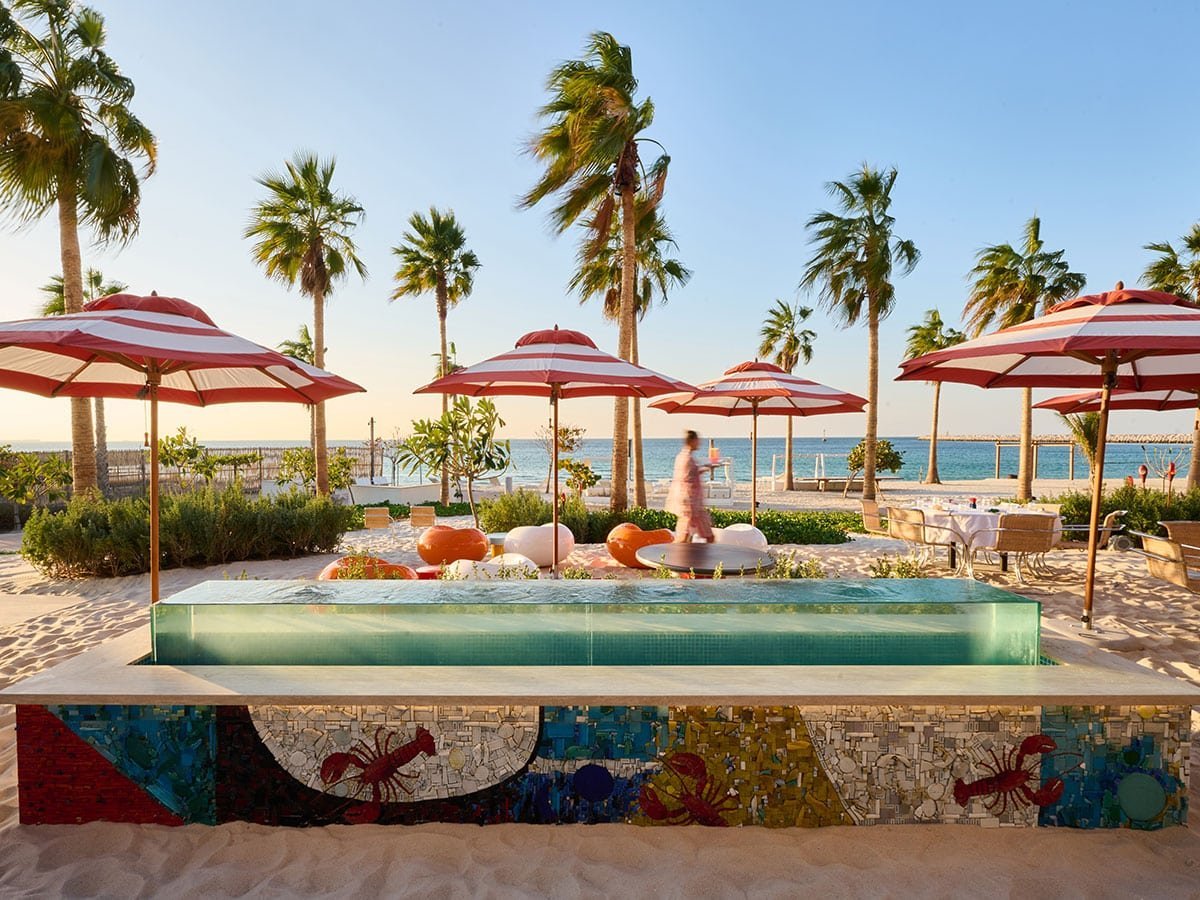 Beachfront pool area with red and white umbrellas, palm trees, and colorful mosaic wall seating, overlooking the ocean