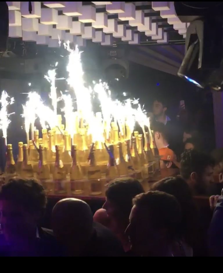 Crowd watching pyrotechnic flames shoot upward from a stage with multiple golden pyro effects during a concert or live performance.
