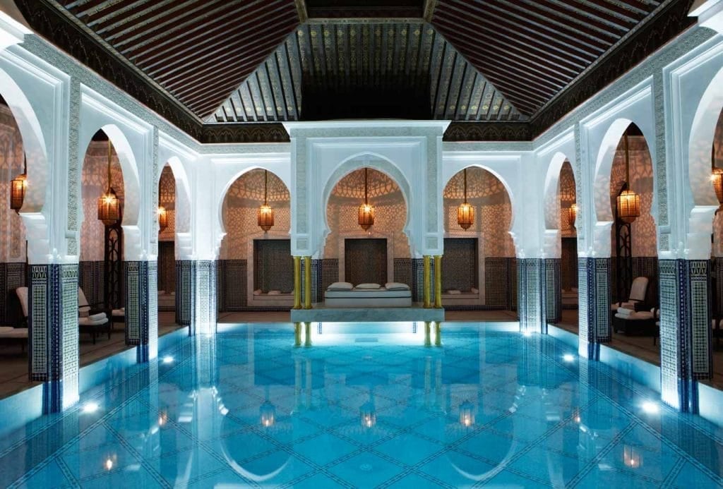 Luxurious indoor swimming pool with blue water, arched doorways, ornate hanging lanterns, and symmetrical Moroccan-style architecture