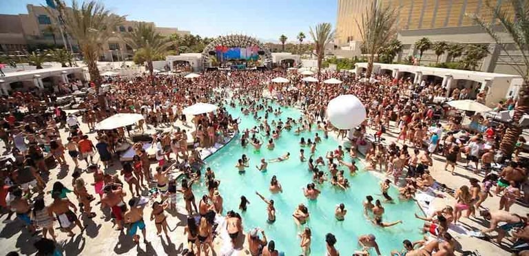 Aerial view of a crowded pool party at a resort with turquoise water, surrounding sun loungers, palm trees, and large crowds of people