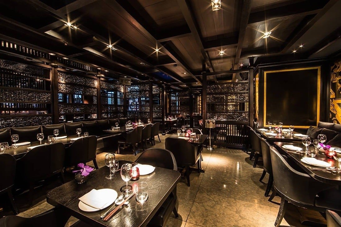 Upscale fine dining restaurant interior with dark wood beams, elegant place settings, purple flowers, and ambient lighting throughout the sophisticated space