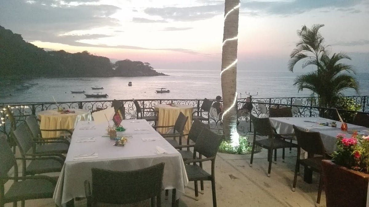 Beachfront restaurant terrace at sunset with white-clothed tables, palm trees, and boats anchored in calm coastal waters