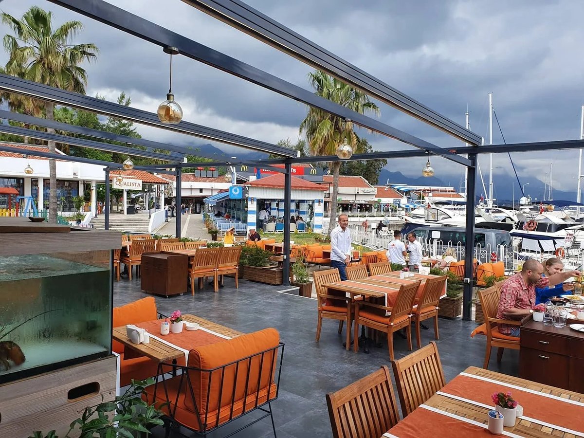 Waterfront restaurant terrace with orange furniture, black steel beams overhead, palm trees, and sailboats in background marina