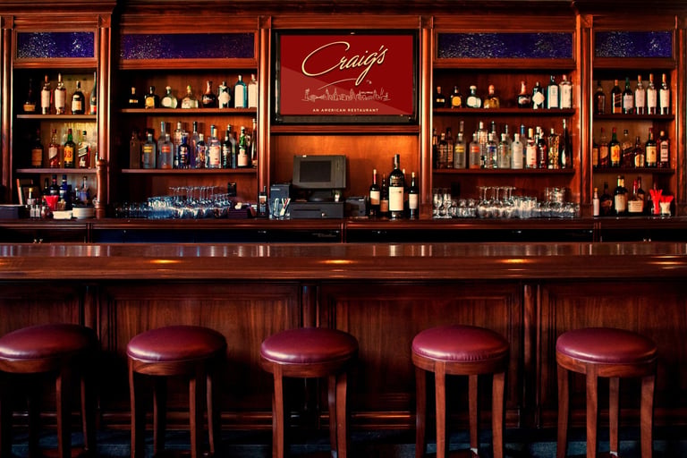 Elegant bar counter with burgundy leather stools, illuminated wooden shelves displaying bottles, and neon signage in a upscale lounge setting