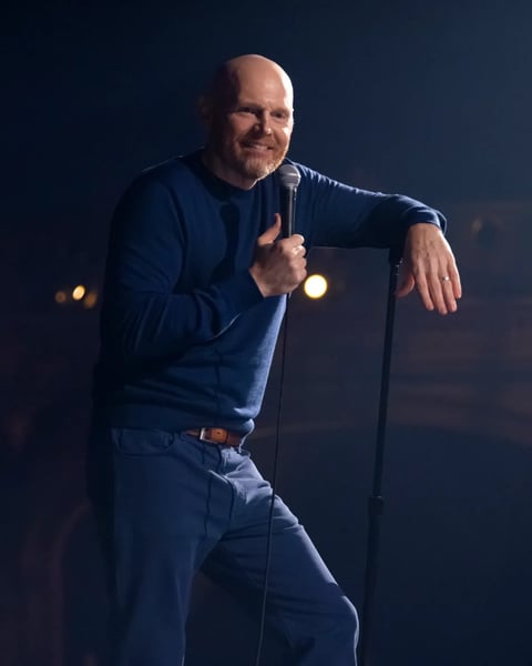 Bald man in blue shirt holding microphone on stage with dark background and stage lights