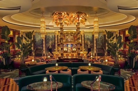 Luxurious hotel lobby with ornate gold ceiling, curved seating, palm decor, and dramatic murals under warm ambient lighting