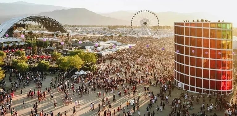 Aerial view of a large outdoor music festival with thousands of attendees, stage structures, and a Ferris wheel against mountain backdrop