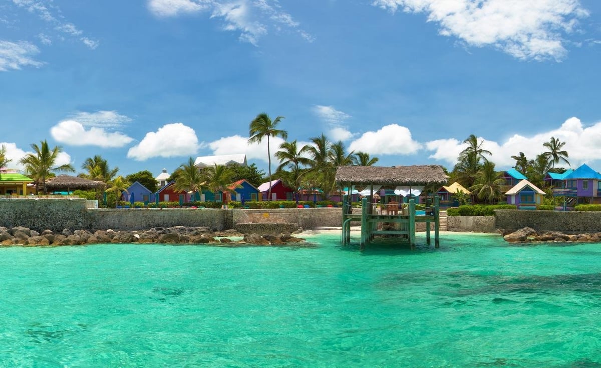Tropical beach village with colorful houses, palm trees, and a green pier extending into turquoise waters under blue sky