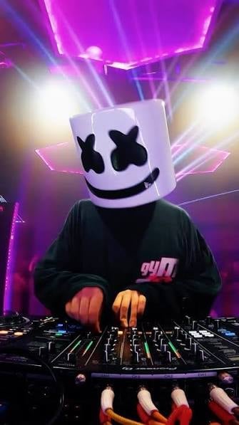 DJ wearing a white marshmallow-shaped helmet with black X eyes and smile, performing at a turntable with purple stage lighting in background