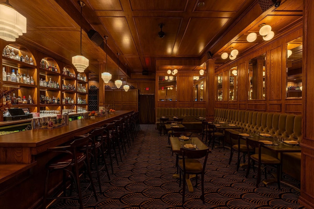 Upscale wood-paneled bar interior with long counter, shelved spirits display, dining tables, and ambient pendant lighting