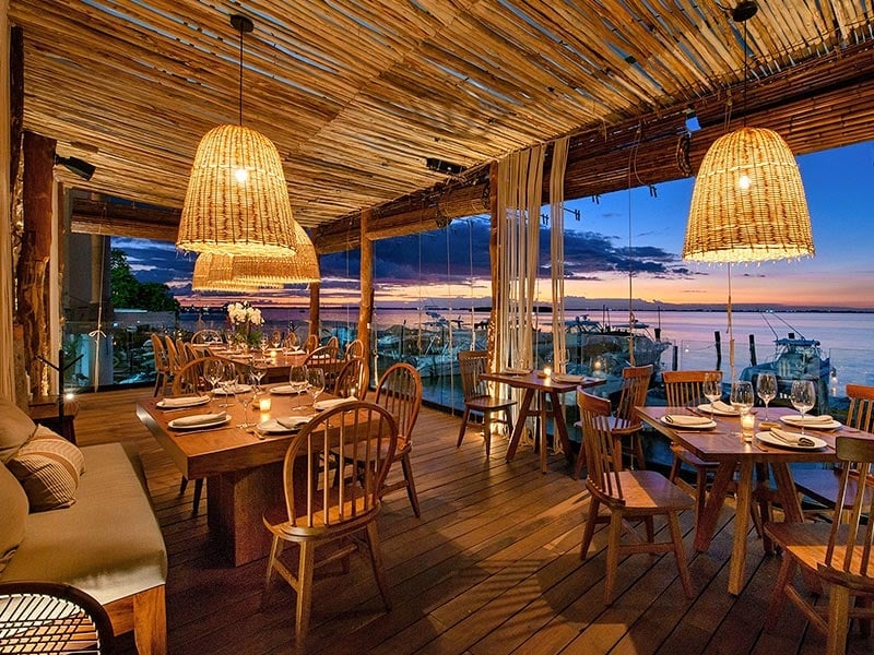 Elegant waterfront restaurant with wooden tables, rattan pendant lights, and panoramic sunset bay views at dusk