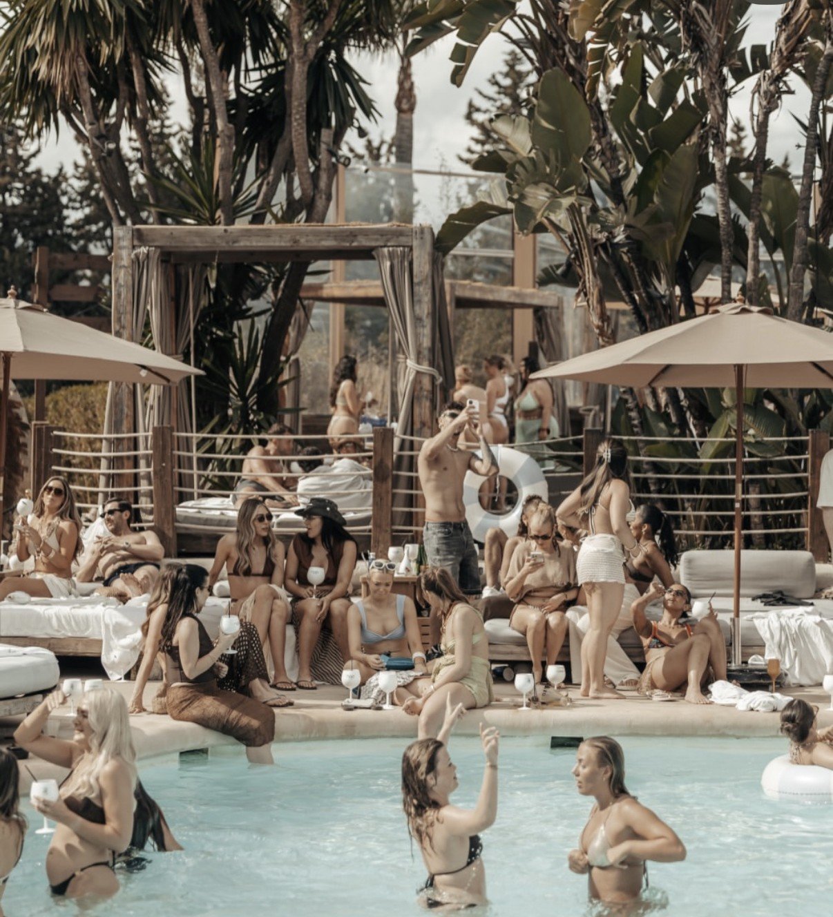 Crowded resort pool scene with people swimming and lounging in cabanas, palm trees overhead, tropical vacation setting
