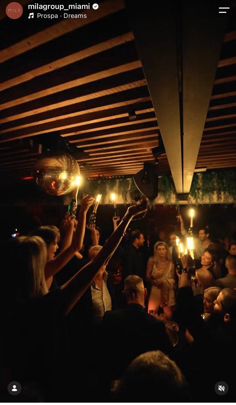 Crowd at outdoor nighttime event under wooden pergola with warm lighting, people with raised arms enjoying music or performance