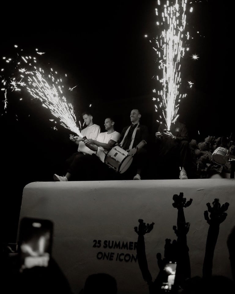 Black and white photo of three men on stage with champagne fountains spraying, silhouettes of audience members below with raised arms.