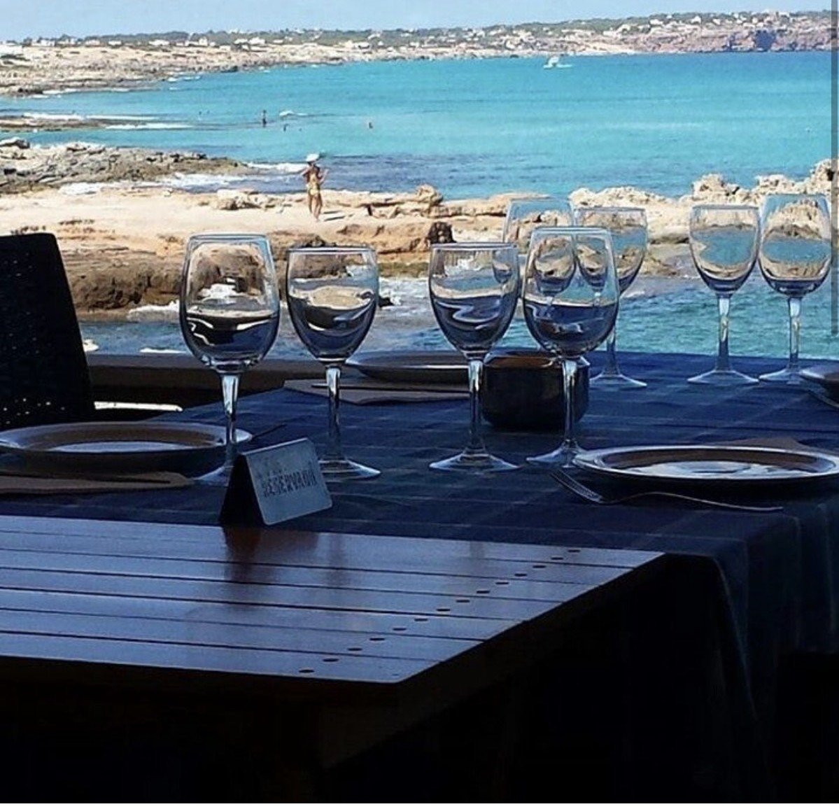 Outdoor dining table with wine glasses overlooking a turquoise Mediterranean sea and sandy beach